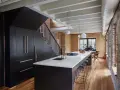 Cucina moderna con isola centrale, bancone in marmo e sgabelli. Armadi neri sotto la scala, con maniglie in legno. Soffitto con travi a vista e tubi. Pareti in mattoni a vista. La **taverna** ha un'**altezza** di circa **2,40** metri.