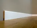 Un battiscopa sagomato bianco, con un sottile bordo in legno a vista, corre lungo una parete grigia e un pavimento in parquet lucido.