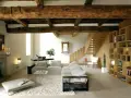 Soggiorno con soffitto con travi in legno a vista, camino in pietra, divano, libreria e scala in legno.