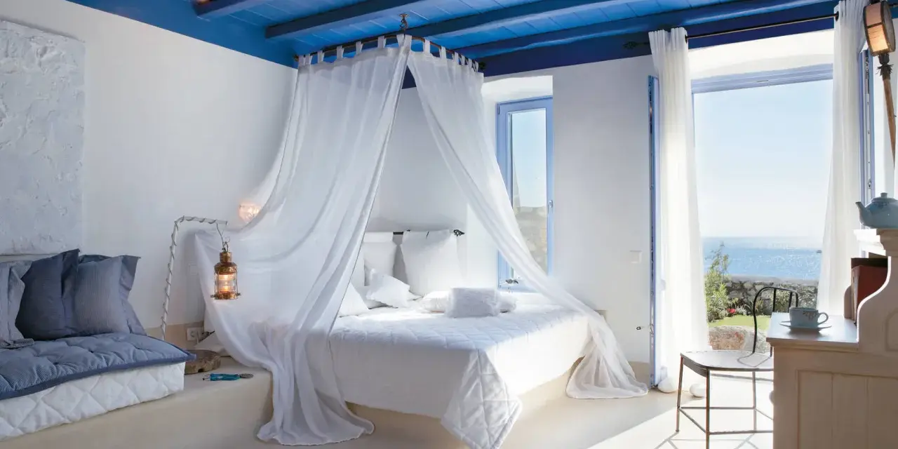 Camera da letto stile mediterraneo con soffitto blu, letto a baldacchino bianco e vista mare.