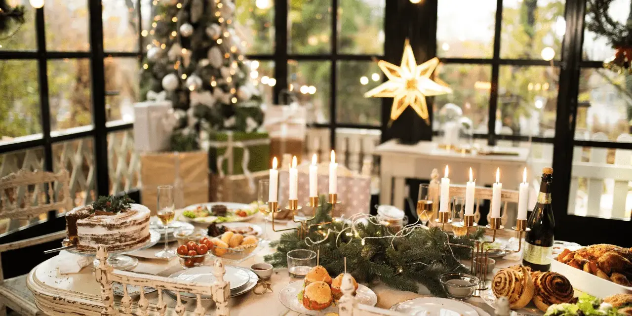 Un tavolo imbandito per Natale, con torta, arrosti e candele. Un modo perfetto per come addobbare la cucina per Natale, con un albero illuminato sullo sfondo.