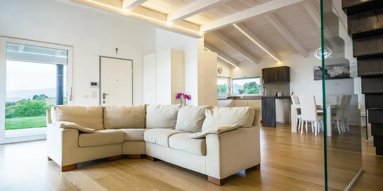 Soggiorno moderno con travi in legno sbiancate senza carteggiare, divano beige e cucina a vista.