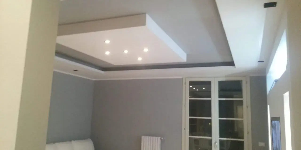 Soffitto moderno con velette in cartongesso, faretti incassati e illuminazione indiretta. Divano bianco e porta finestra.