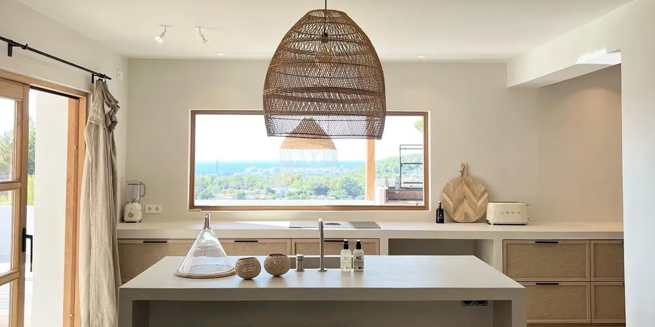 Cucina luminosa con isola centrale, lampadario in rattan e vista mare. Arredamento in stile casa bohemien con materiali naturali.
