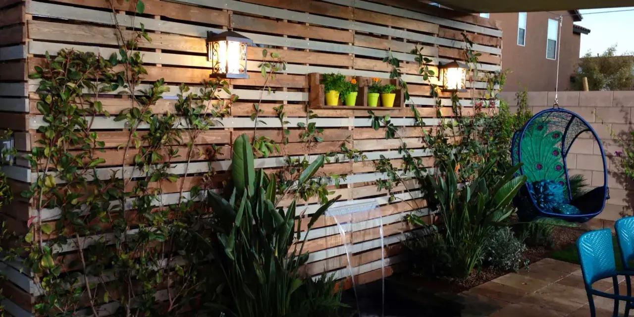 Giardino incantevole con idee creative con bancali: una parete in legno, cascata, piante rigogliose e una sedia sospesa a forma di pavone.