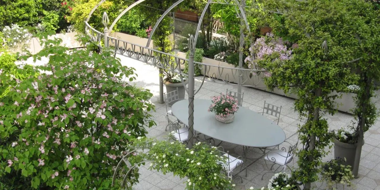 Un angolo incantevole per idee giardino villa: un pergolato metallico abbraccia un tavolo ovale con sedie, circondato da fioriture rosa e verdi rigogliosi.