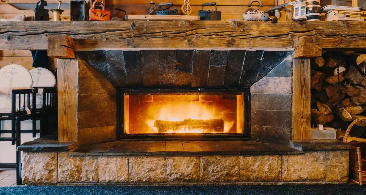 Camino in pietra e legno, con fuoco acceso, che evoca idee camini rustici di montagna. Sopra, oggetti d'epoca e legna accatastata.