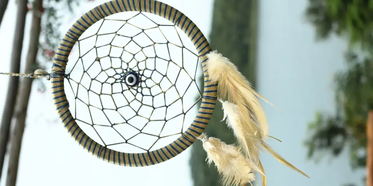 Acchiappasogni fai da te con perline e piume, decorato con un occhio blu al centro, appeso tra alberi e fiori.