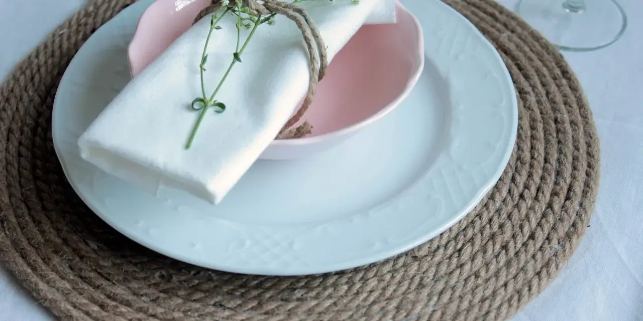 Sottopiatti in stoffa fai da te, con tovagliolo bianco legato da spago e decorato con un rametto fiorito.