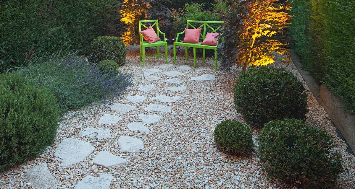 Giardino pavimentato con aiuole di lavanda e cespugli. Due sedie verdi con cuscini rosa invitano al relax.
