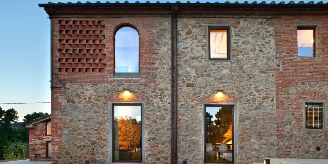 Esterno di case stile toscano con muri in pietra e mattoni, finestre ad arco e rettangolari, illuminazione calda.