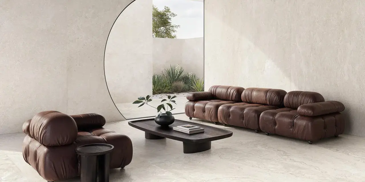 Salotto moderno con divano e poltrona in pelle marrone, tavolino in legno scuro e vista su un giardino interno. L'ambiente evoca la serenità di una casa provenzale.