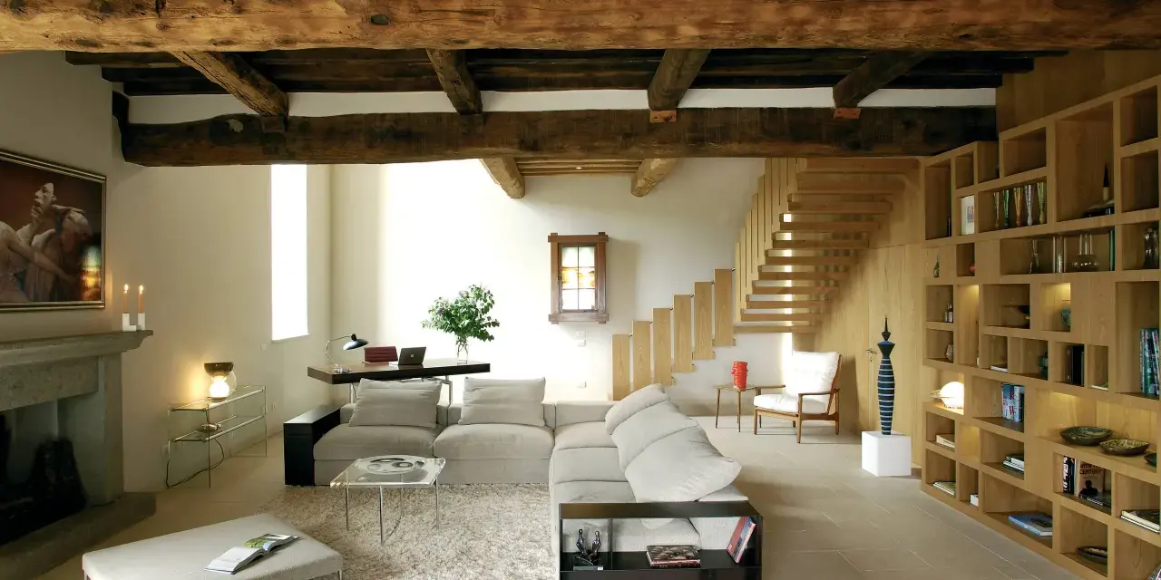 Soggiorno con soffitto con travi in legno a vista, camino in pietra, divano, libreria e scala in legno.
