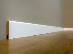 Un battiscopa sagomato bianco, con un sottile bordo in legno a vista, corre lungo una parete grigia e un pavimento in parquet lucido.