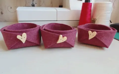Tre cestini in feltro fai da te, color melanzana, decorati con bottoni a forma di cuore. Perfetti per organizzare piccoli oggetti.