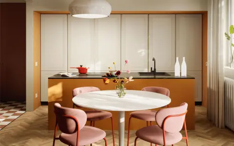 Una casa colorata con un'isola cucina arancione, sedie rosa e un tavolo bianco. Un vaso di fiori aggiunge un tocco di vivacità.