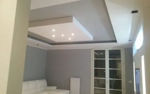 Soffitto moderno con velette in cartongesso, faretti incassati e illuminazione indiretta. Divano bianco e porta finestra.