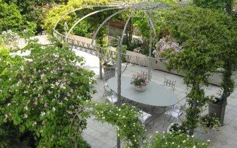 Un angolo incantevole per idee giardino villa: un pergolato metallico abbraccia un tavolo ovale con sedie, circondato da fioriture rosa e verdi rigogliosi.