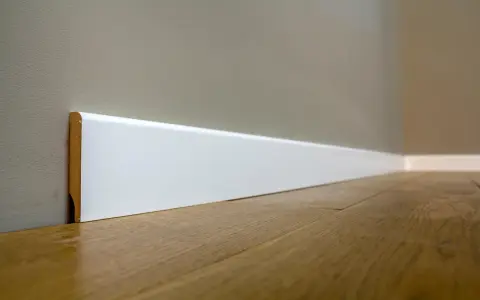 Un battiscopa sagomato bianco, con un sottile bordo in legno a vista, corre lungo una parete grigia e un pavimento in parquet lucido.