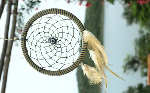 Acchiappasogni fai da te con perline e piume, decorato con un occhio blu al centro, appeso tra alberi e fiori.