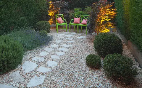 Giardino pavimentato con aiuole di lavanda e cespugli. Due sedie verdi con cuscini rosa invitano al relax.