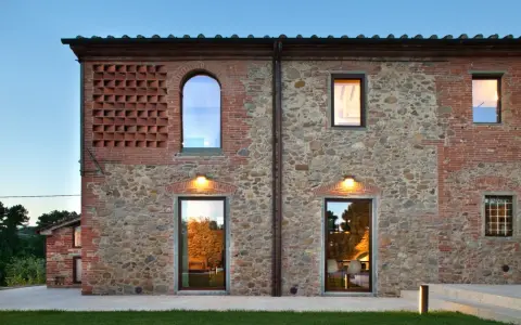 Esterno di case stile toscano con muri in pietra e mattoni, finestre ad arco e rettangolari, illuminazione calda.