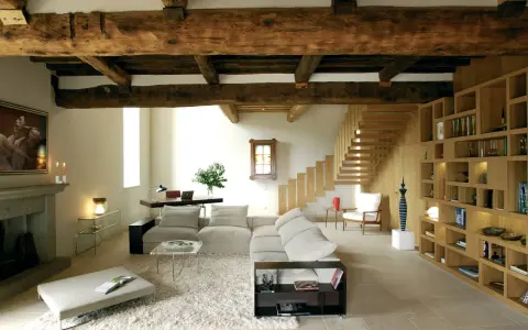 Soggiorno con soffitto con travi in legno a vista, camino in pietra, divano, libreria e scala in legno.
