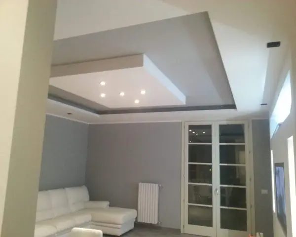 Soffitto moderno con velette in cartongesso, faretti incassati e illuminazione indiretta. Divano bianco e porta finestra.