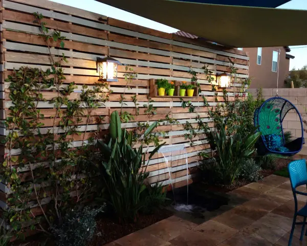 Giardino incantevole con idee creative con bancali: una parete in legno, cascata, piante rigogliose e una sedia sospesa a forma di pavone.