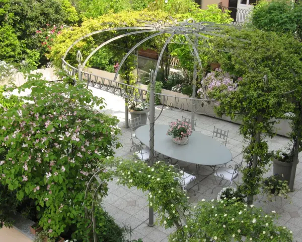 Un angolo incantevole per idee giardino villa: un pergolato metallico abbraccia un tavolo ovale con sedie, circondato da fioriture rosa e verdi rigogliosi.