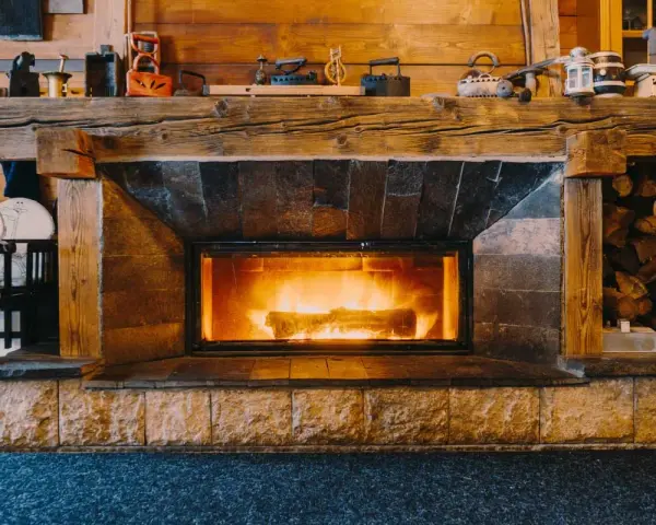 Camino in pietra e legno, con fuoco acceso, che evoca idee camini rustici di montagna. Sopra, oggetti d'epoca e legna accatastata.
