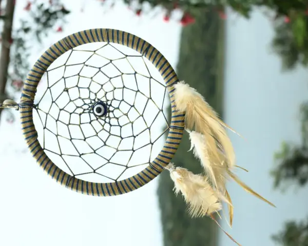 Acchiappasogni fai da te con perline e piume, decorato con un occhio blu al centro, appeso tra alberi e fiori.