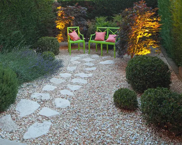 Giardino pavimentato con aiuole di lavanda e cespugli. Due sedie verdi con cuscini rosa invitano al relax.