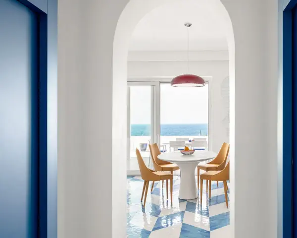 Interno di una casa stile mare con pavimento a scacchi blu e bianchi, tavolo rotondo con sedie arancioni e vista sul mare.