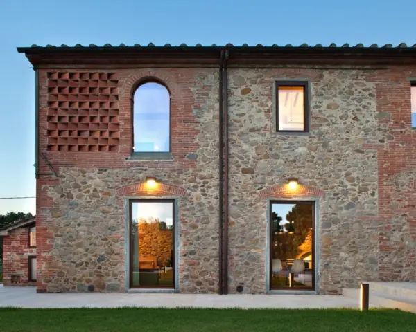 Esterno di case stile toscano con muri in pietra e mattoni, finestre ad arco e rettangolari, illuminazione calda.