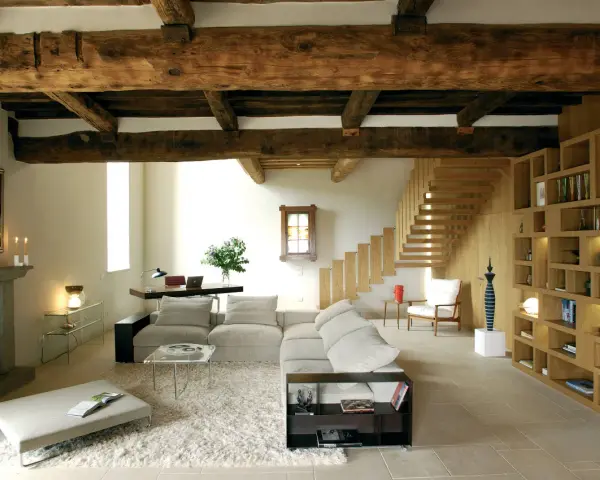 Soggiorno con soffitto con travi in legno a vista, camino in pietra, divano, libreria e scala in legno.