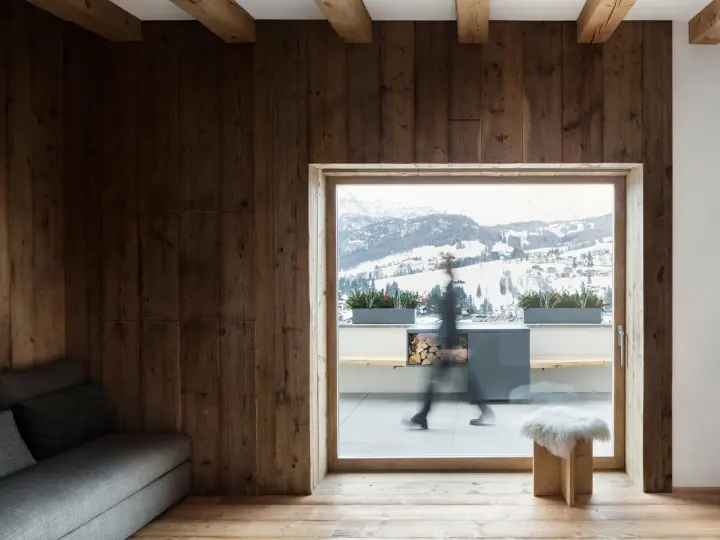 Interni di case di montagna con pareti in legno scuro, travi a vista e un divano grigio. Fuori, un paesaggio innevato.