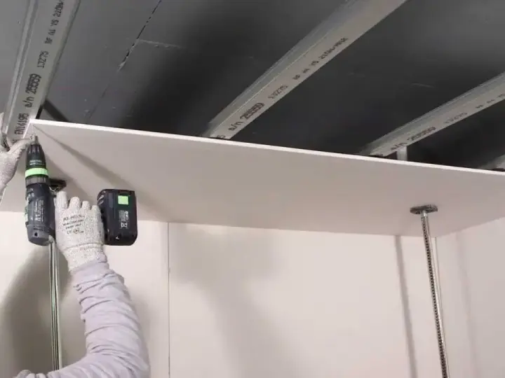 Persona con guanti avvita una lastra di cartongesso al soffitto metallico, un progetto di cartongesso fai da te.