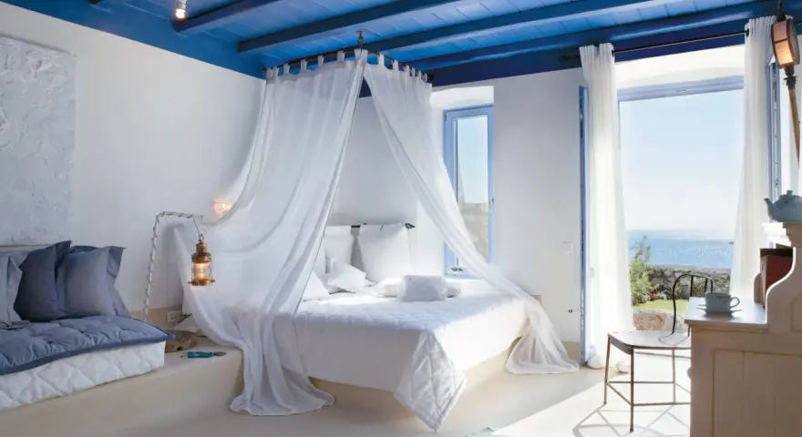 Camera da letto stile mediterraneo con soffitto blu, letto a baldacchino bianco e vista mare.