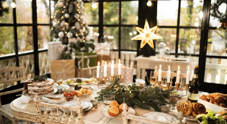 Un tavolo imbandito per Natale, con torta, arrosti e candele. Un modo perfetto per come addobbare la cucina per Natale, con un albero illuminato sullo sfondo.