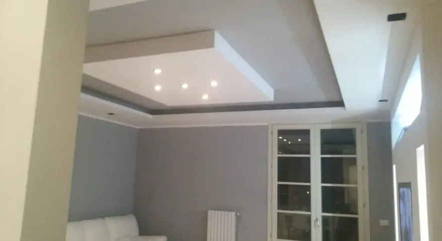 Soffitto moderno con velette in cartongesso, faretti incassati e illuminazione indiretta. Divano bianco e porta finestra.