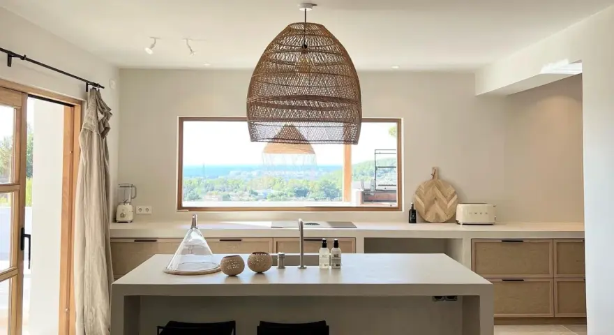 Cucina luminosa con isola centrale, lampadario in rattan e vista mare. Arredamento in stile casa bohemien con materiali naturali.