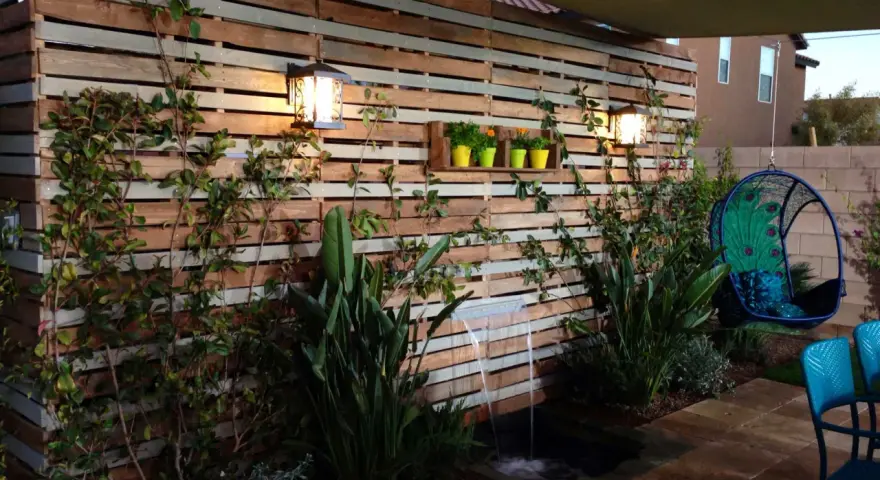Giardino incantevole con idee creative con bancali: una parete in legno, cascata, piante rigogliose e una sedia sospesa a forma di pavone.