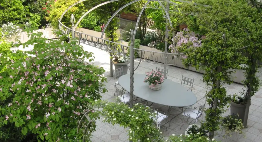 Un angolo incantevole per idee giardino villa: un pergolato metallico abbraccia un tavolo ovale con sedie, circondato da fioriture rosa e verdi rigogliosi.
