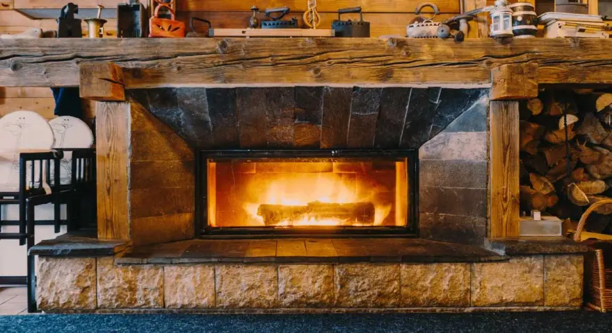 Camino in pietra e legno, con fuoco acceso, che evoca idee camini rustici di montagna. Sopra, oggetti d'epoca e legna accatastata.