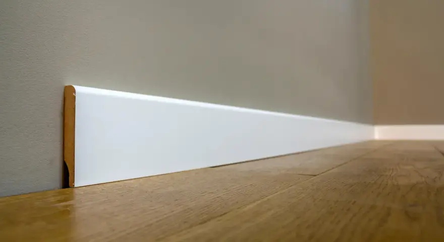 Un battiscopa sagomato bianco, con un sottile bordo in legno a vista, corre lungo una parete grigia e un pavimento in parquet lucido.