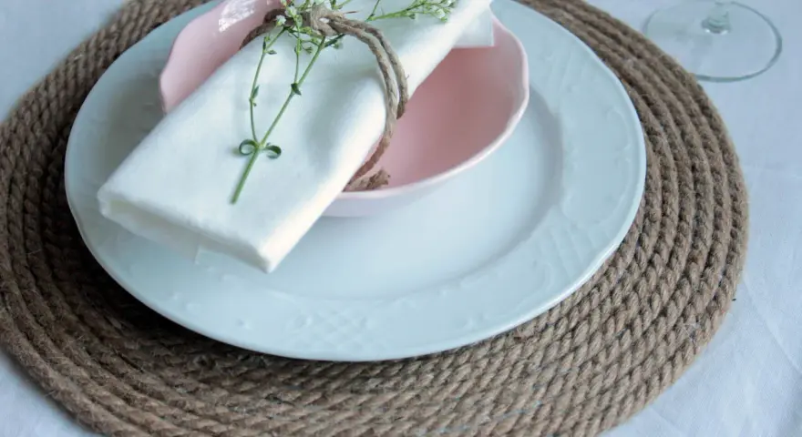 Sottopiatti in stoffa fai da te, con tovagliolo bianco legato da spago e decorato con un rametto fiorito.
