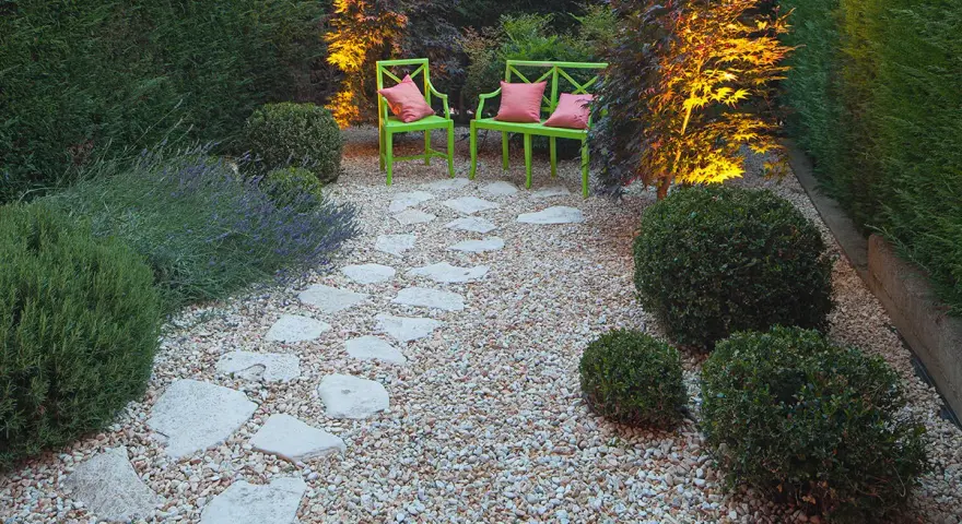Giardino pavimentato con aiuole di lavanda e cespugli. Due sedie verdi con cuscini rosa invitano al relax.