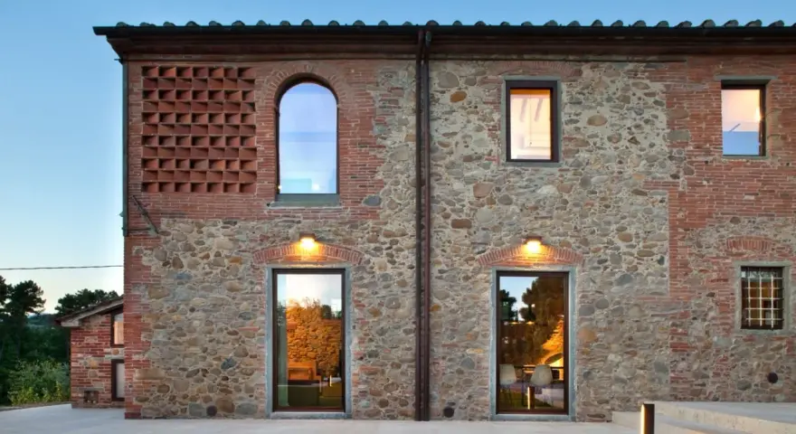 Esterno di case stile toscano con muri in pietra e mattoni, finestre ad arco e rettangolari, illuminazione calda.