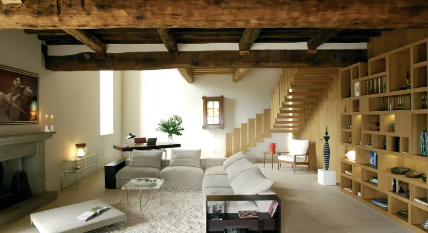 Soggiorno con soffitto con travi in legno a vista, camino in pietra, divano, libreria e scala in legno.